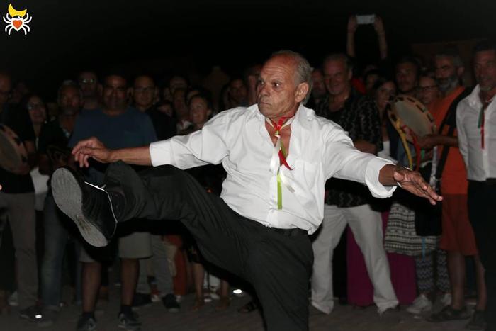 Danzatore con una gamba sollevata al centro della ronda notturna, circondato dal pubblico e da suonatori di tamburello.